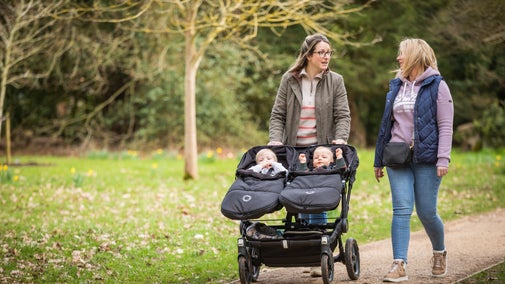 Two women walking down a path. One is pushing a stroller with two babies in.